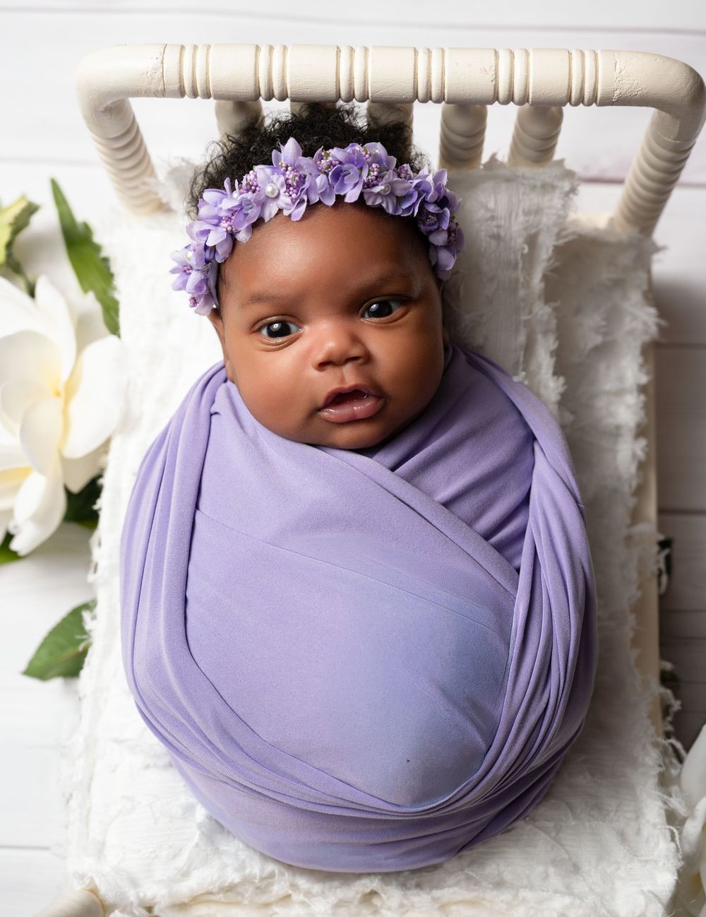 Newborn baby swaddled in lavender wrap with purple floral headband, lying in a small white wooden bed.