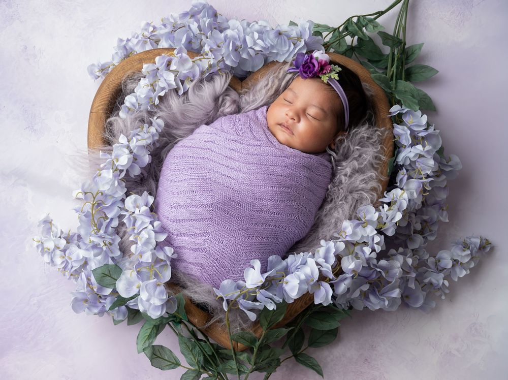 Newborn baby swaddled in lavender wrap sleeping in wooden bowl with purple flowers and floral headband.
