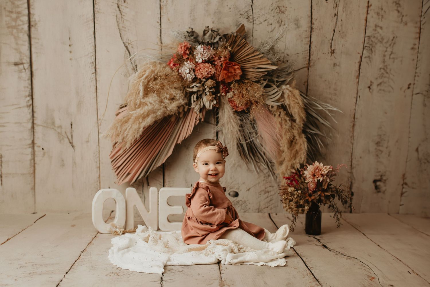 Rustic first birthday setup with dried flower arch and decorative letters against white backdrop.