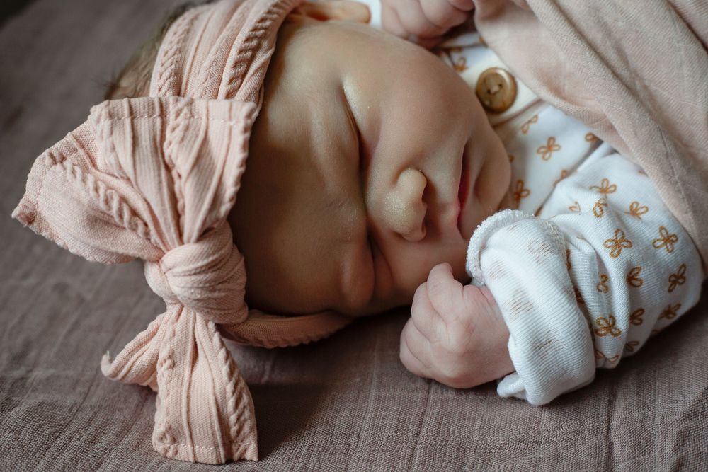 Sleeping infant with pink bow headband wrapped in soft fabric.