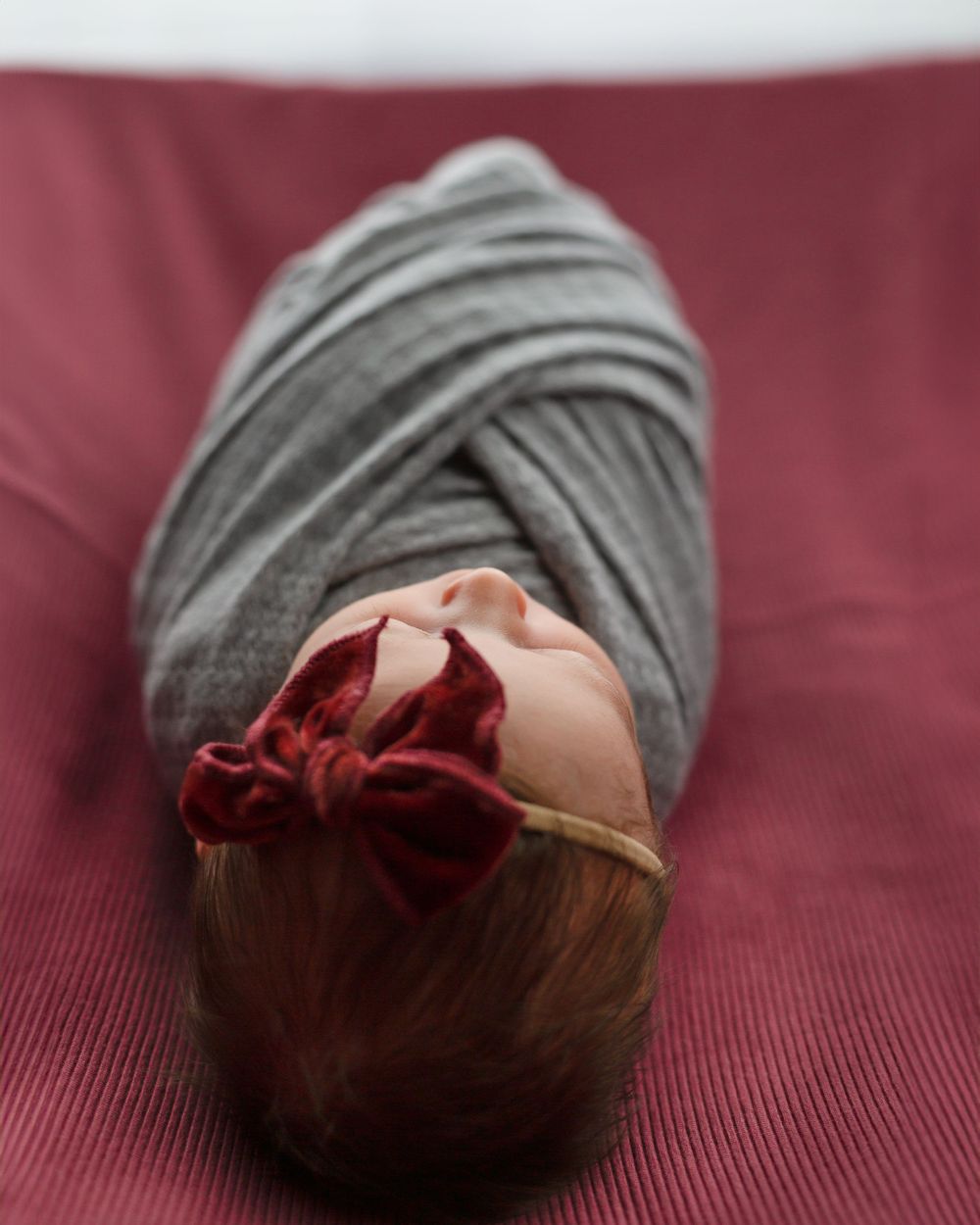 Sleeping newborn wrapped in gray swaddle with red bow on burgundy background.