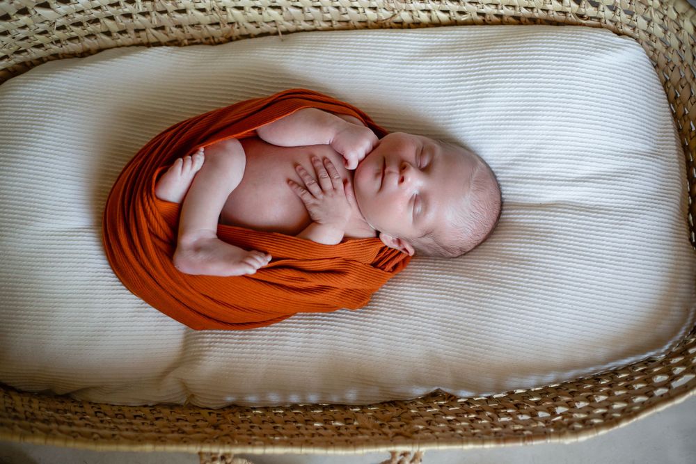 Sleeping newborn wrapped in orange cloth lying in woven bassinet.