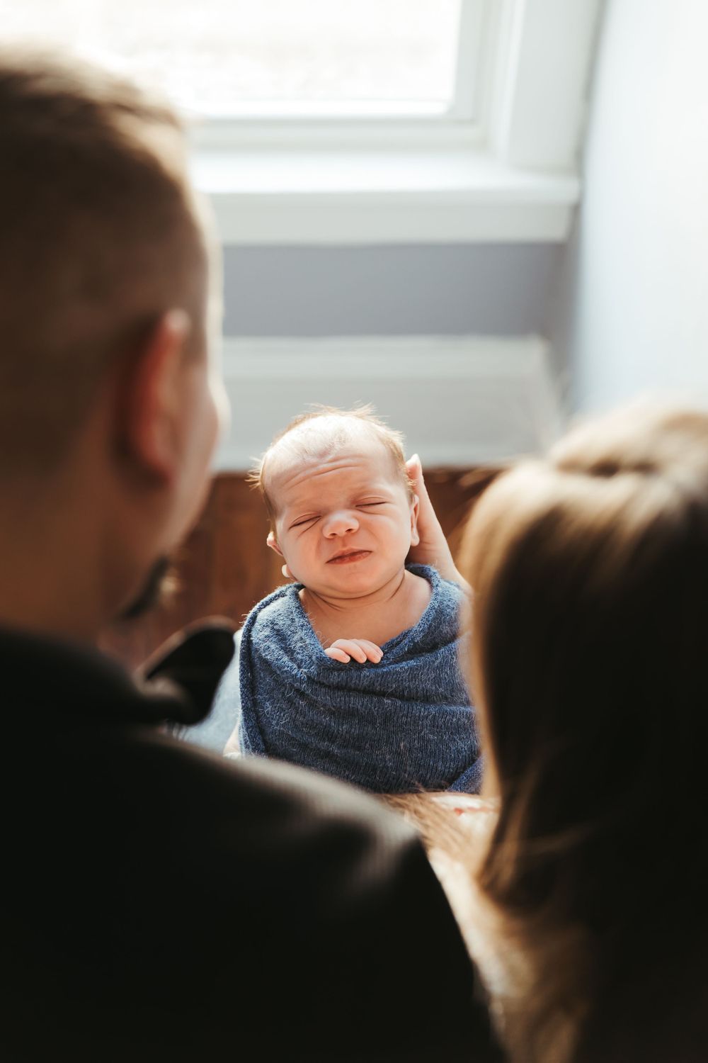 A peaceful resting newborn wrapped in a soft gray blanket while parents look on lovingly in natural window light.