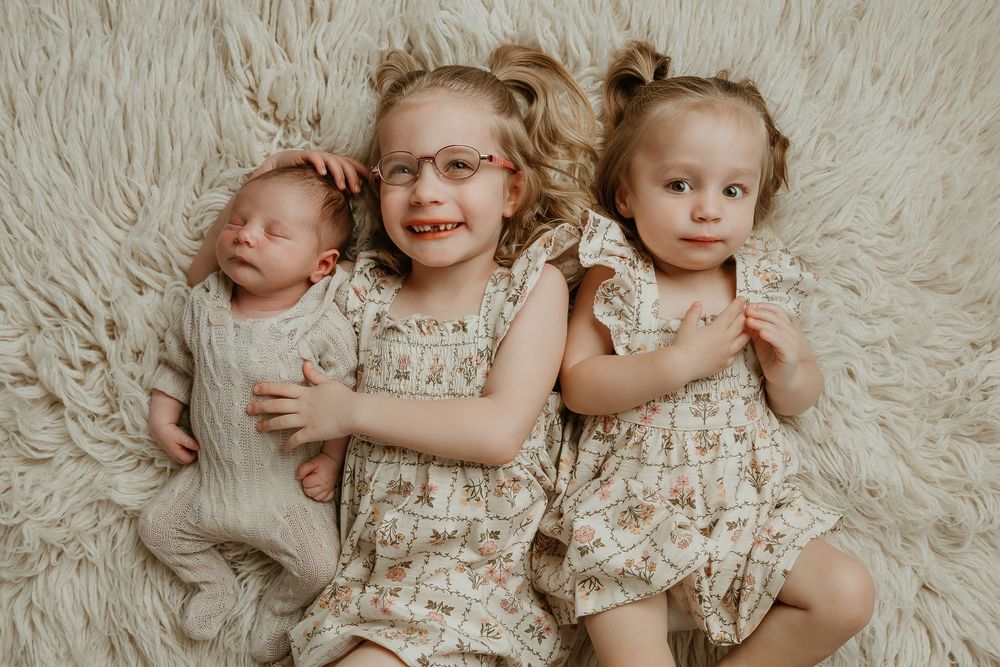Three young sisters lying on a fluffy cream rug, including a newborn baby, toddler, and older girl wearing matching floral dresses.