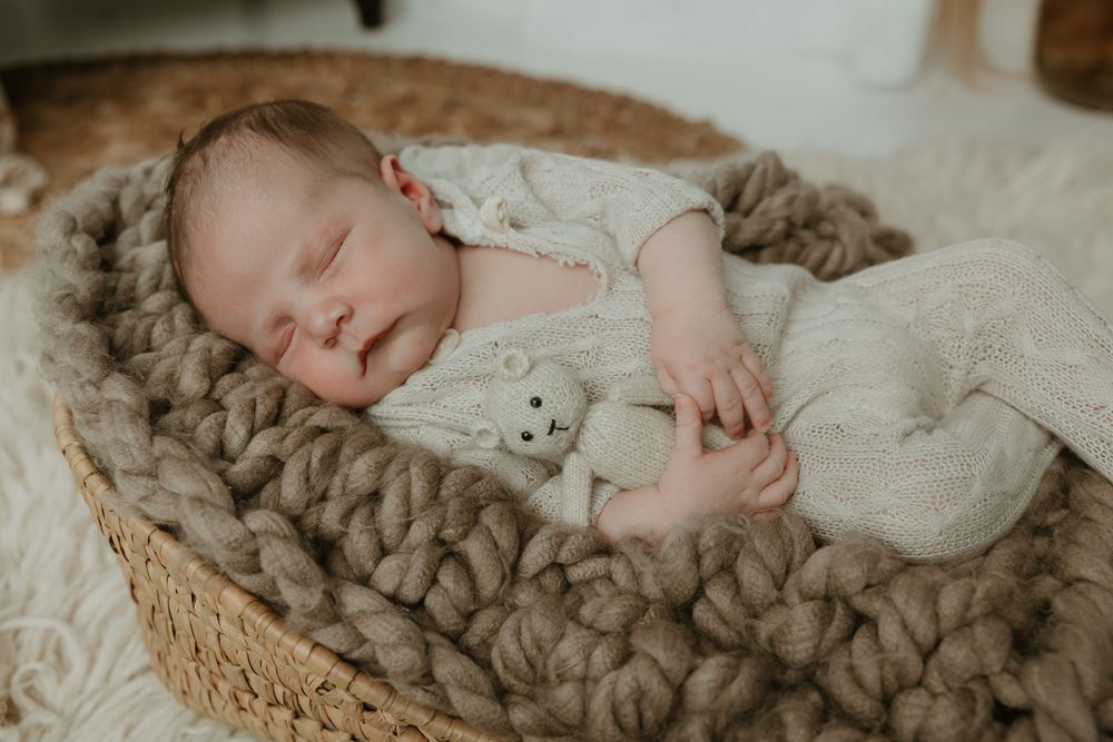 Newborn baby sleeping peacefully in a wicker basket with chunky knit blanket and white stuffed animal toy.