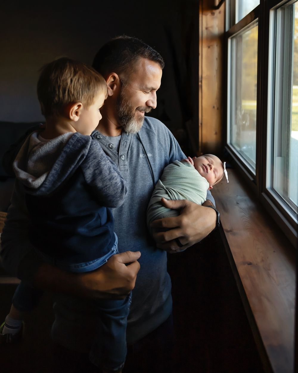 Intimate family moment by window as father holds toddler and newborn.