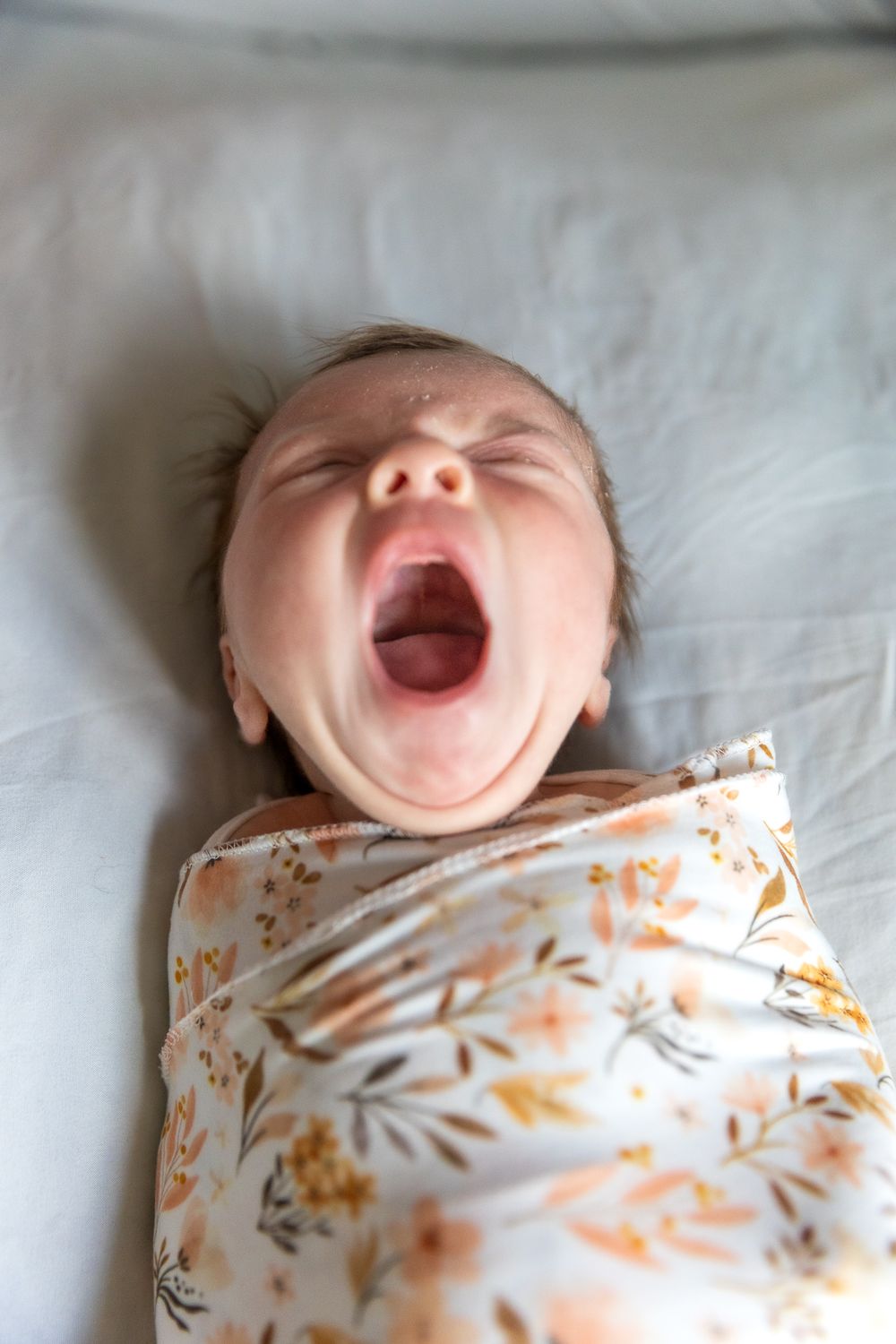 Baby lying on white bedding yawning with mouth wide open wearing floral print outfit.