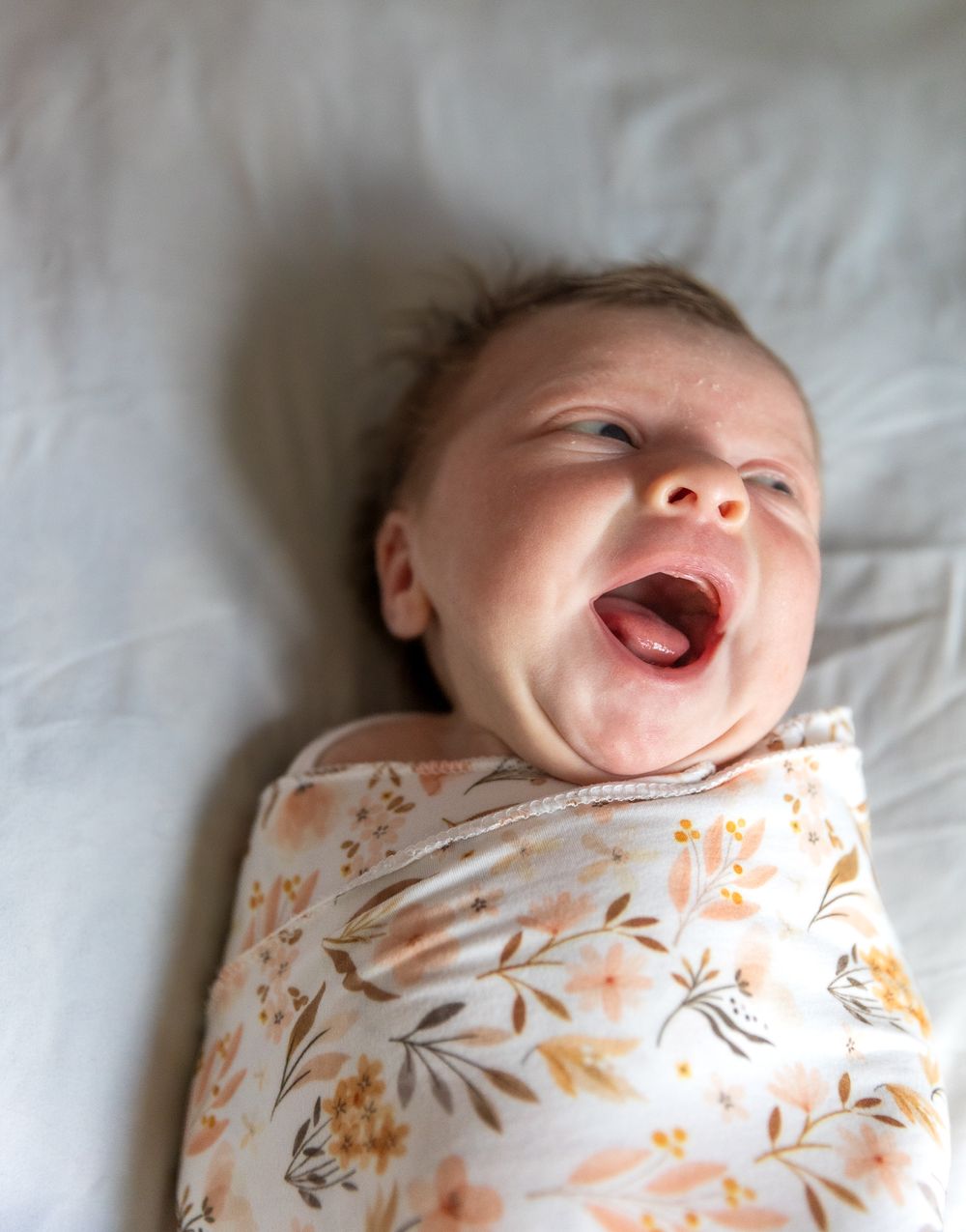 A baby wearing a floral patterned onesie lies on white bedding while laughing joyfully.