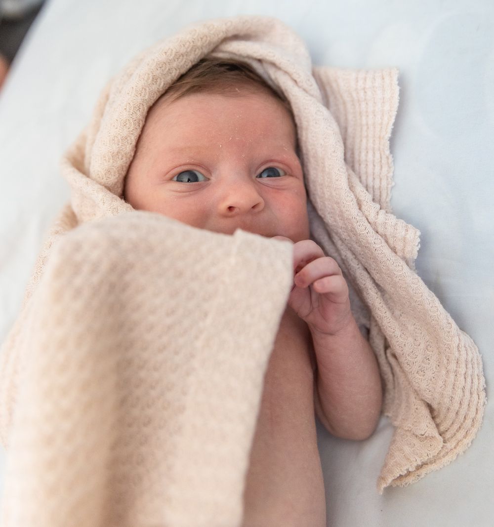 A baby wrapped in a soft beige blanket peeks out with bright eyes.