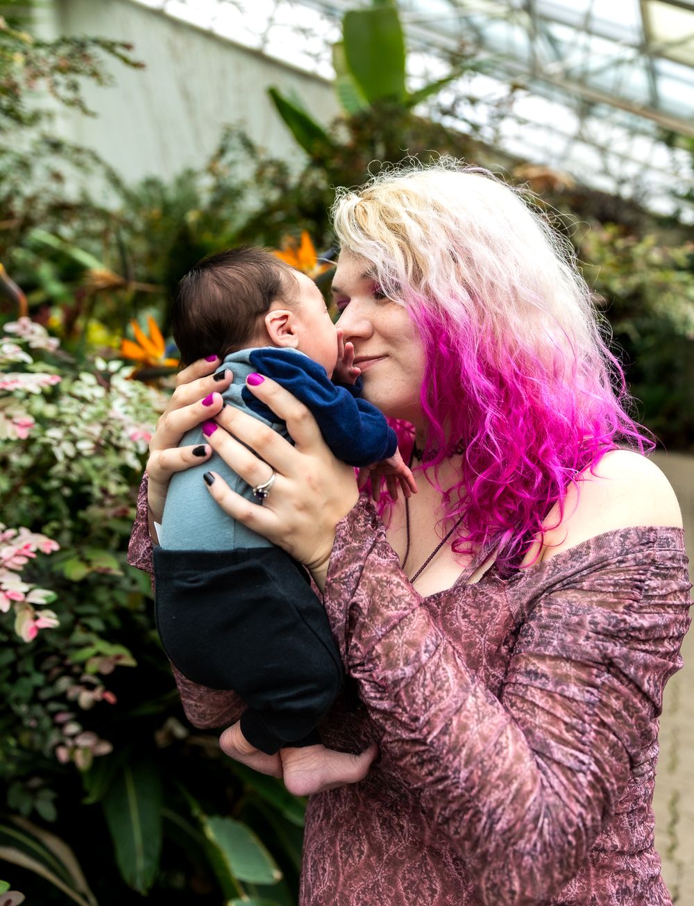Parent with vibrant pink hair holding infant in greenhouse setting surrounded by plants.
