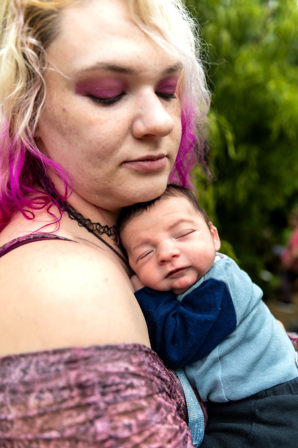 Intimate portrait of parent with pink-tipped hair holding sleeping infant outdoors.
