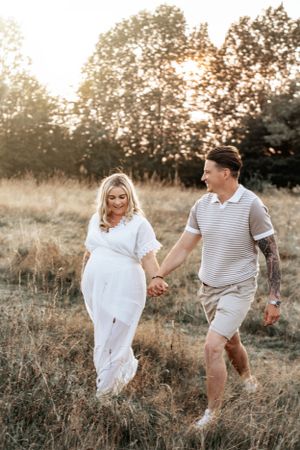Couple walking hand in hand through a golden summer meadow at sunset with trees in background. Maternity Photoshoot.