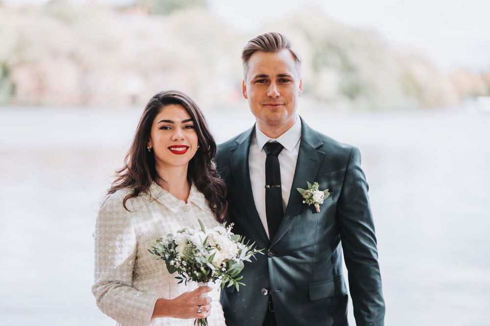 A couple in wedding attire posing outdoors with a waterfront backdrop.
