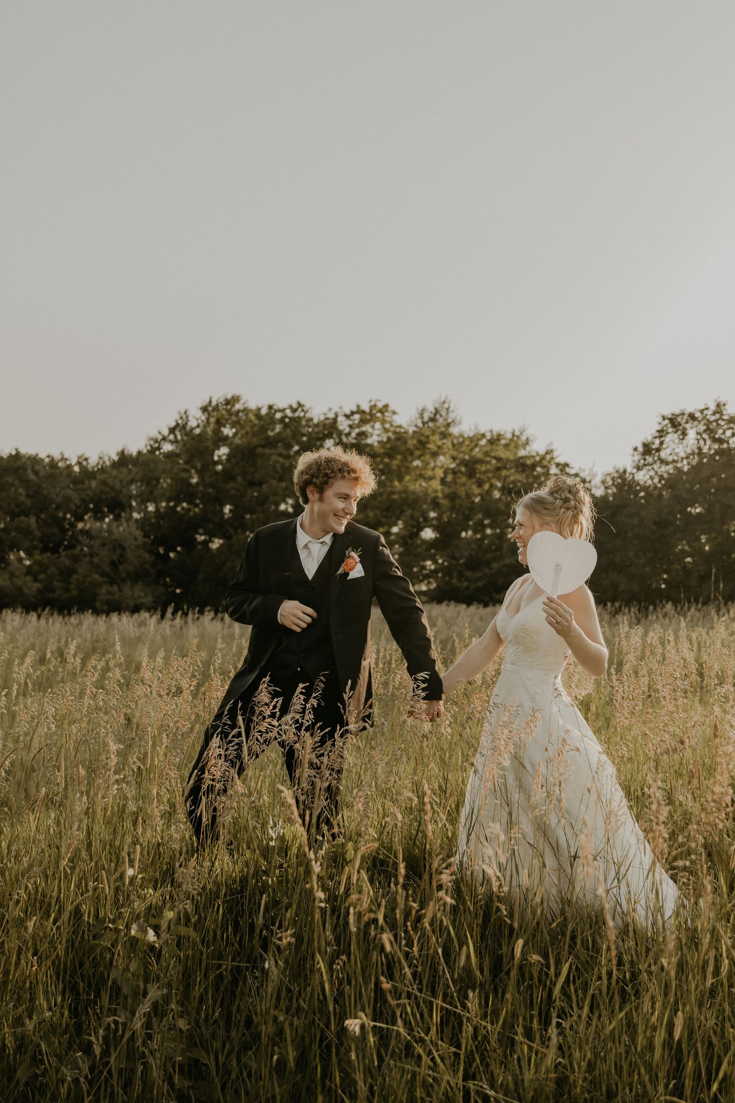 A couple in wedding attire walks through a golden field of tall grass at sunset, with trees in the background.