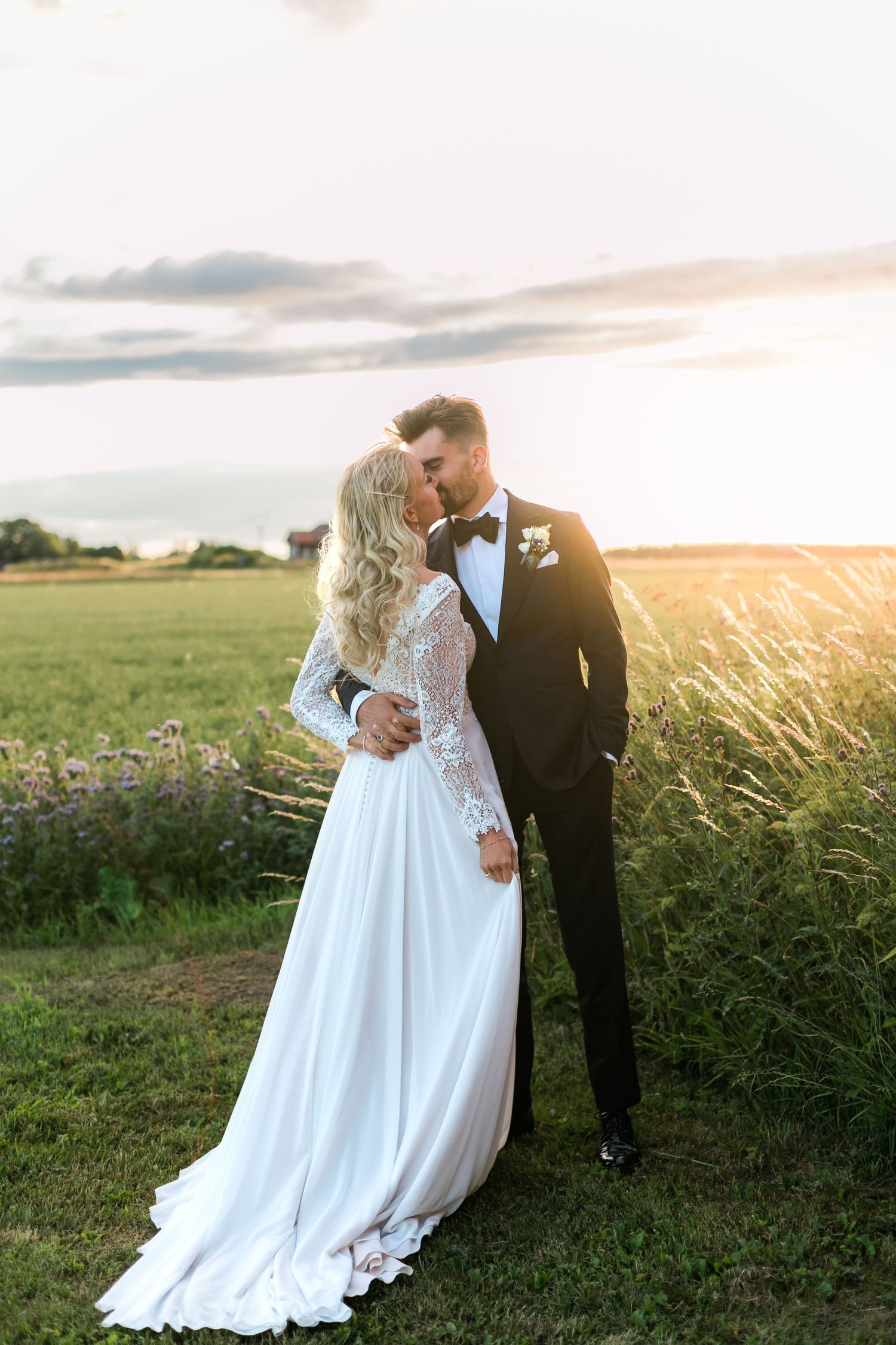Romantic wedding couple embraces in a golden sunlit field at sunset with bride wearing lace-sleeved white gown.