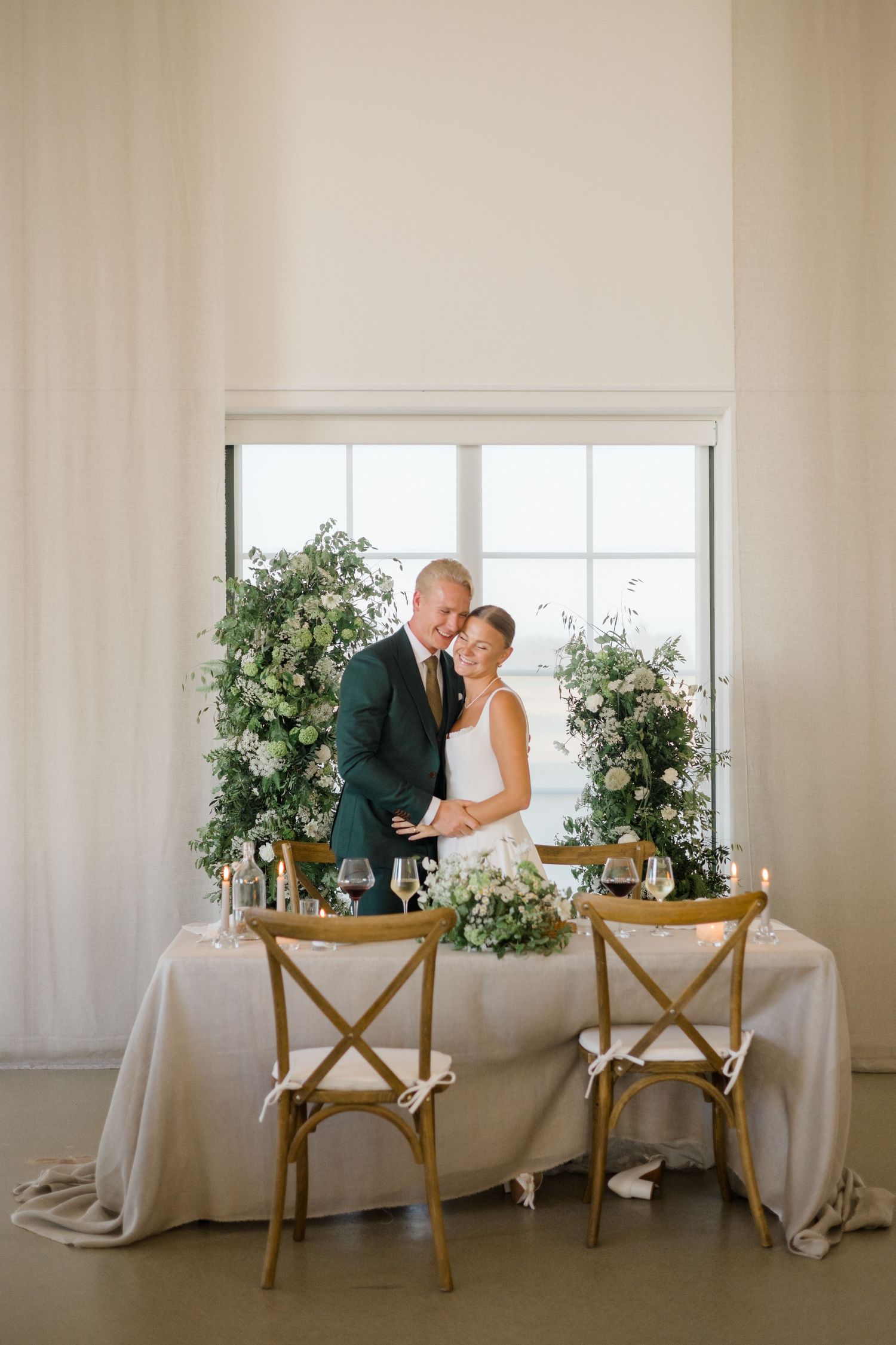 Elegant wedding sweetheart table setup with greenery arrangements and wooden crossback chairs against a bright window.