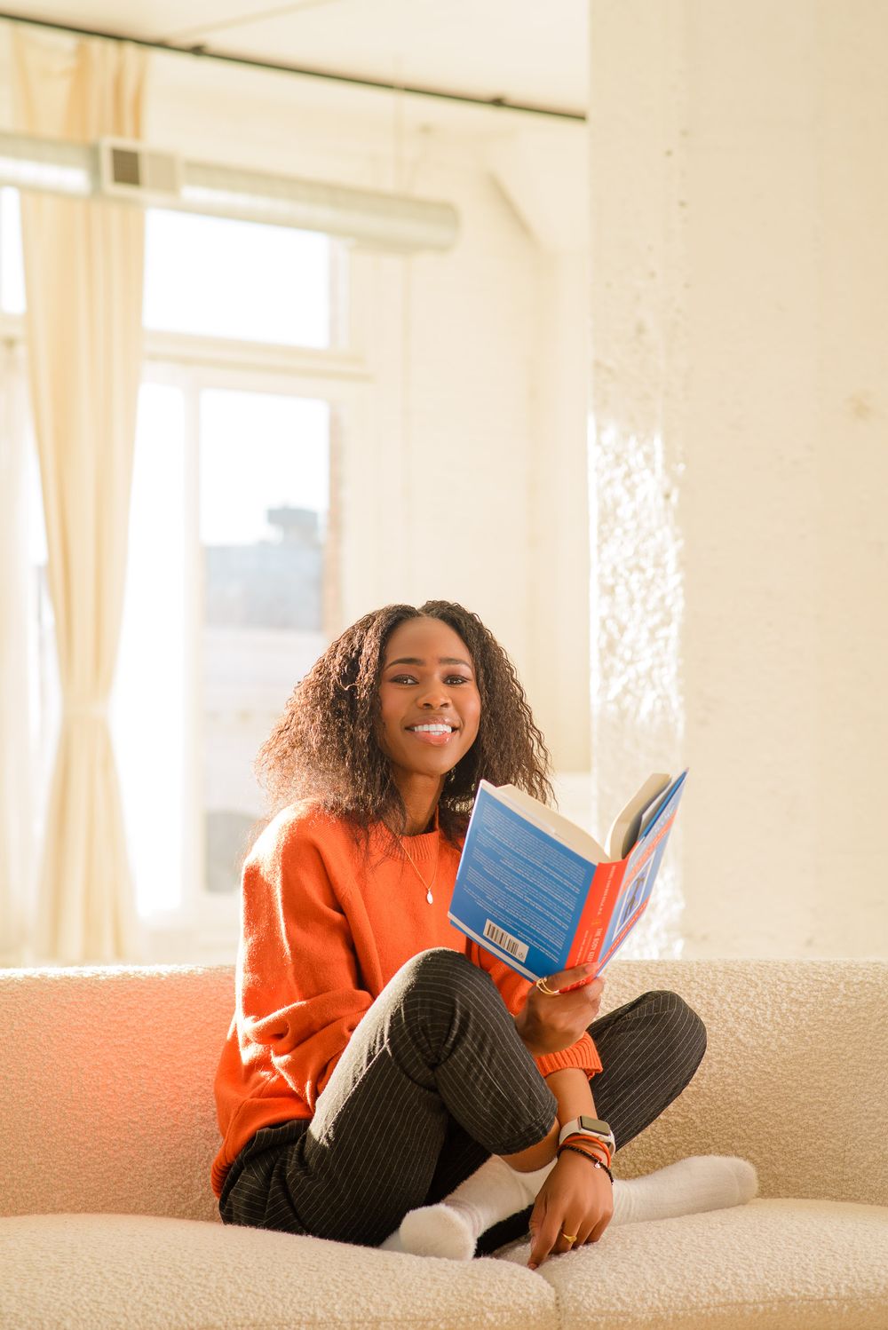 Person in orange sweater relaxing on beige couch with book.