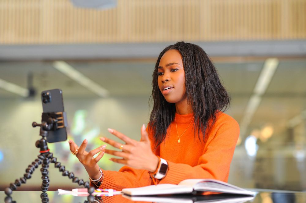 A content creator sitting in a bright room using a mobile device on a tripod to film content.