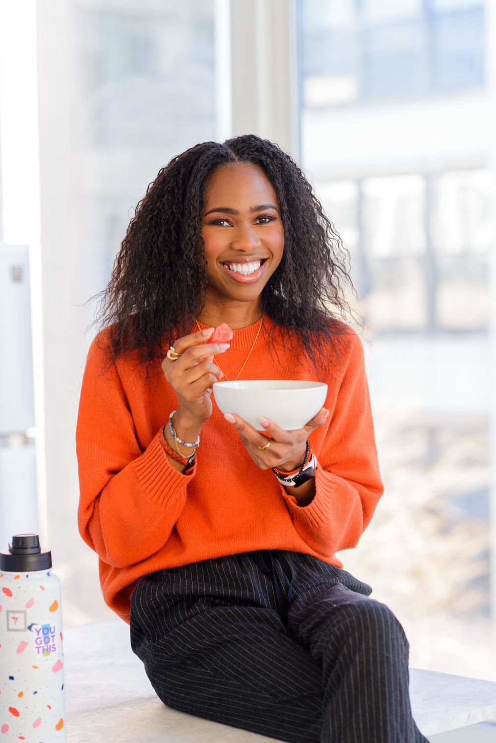 Someone in a bright orange sweater enjoying a meal from a white bowl while relaxing by a window.