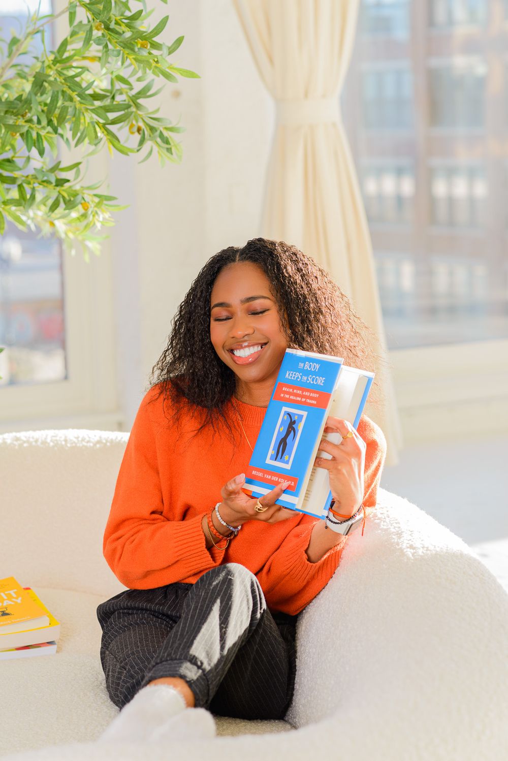 Someone wearing orange relaxing on a white couch with a blue book in a sunlit room with plants.