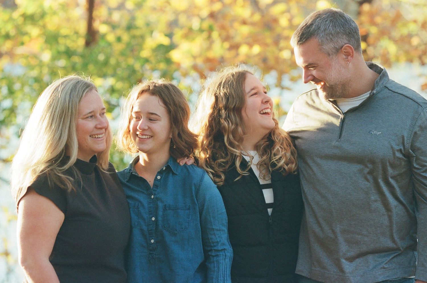 A family wearing casual clothing stands together outdoors during autumn with golden foliage in the background.