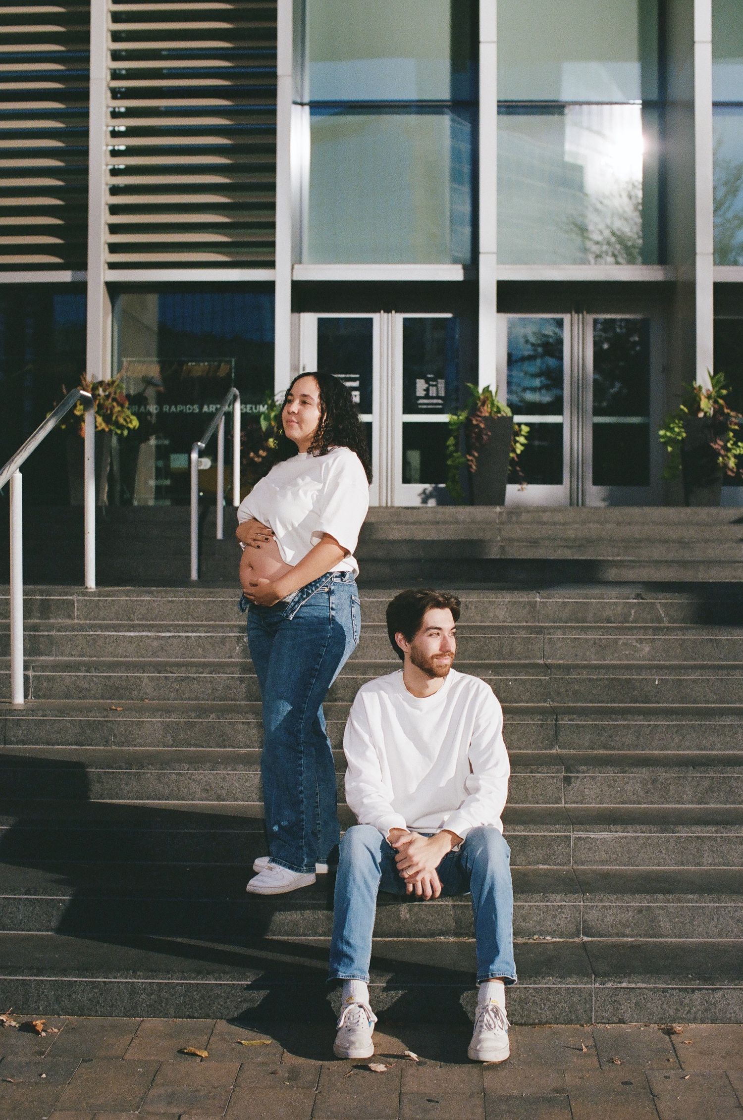 Two people in casual white t-shirts and jeans sit on concrete steps outside a modern building entrance.