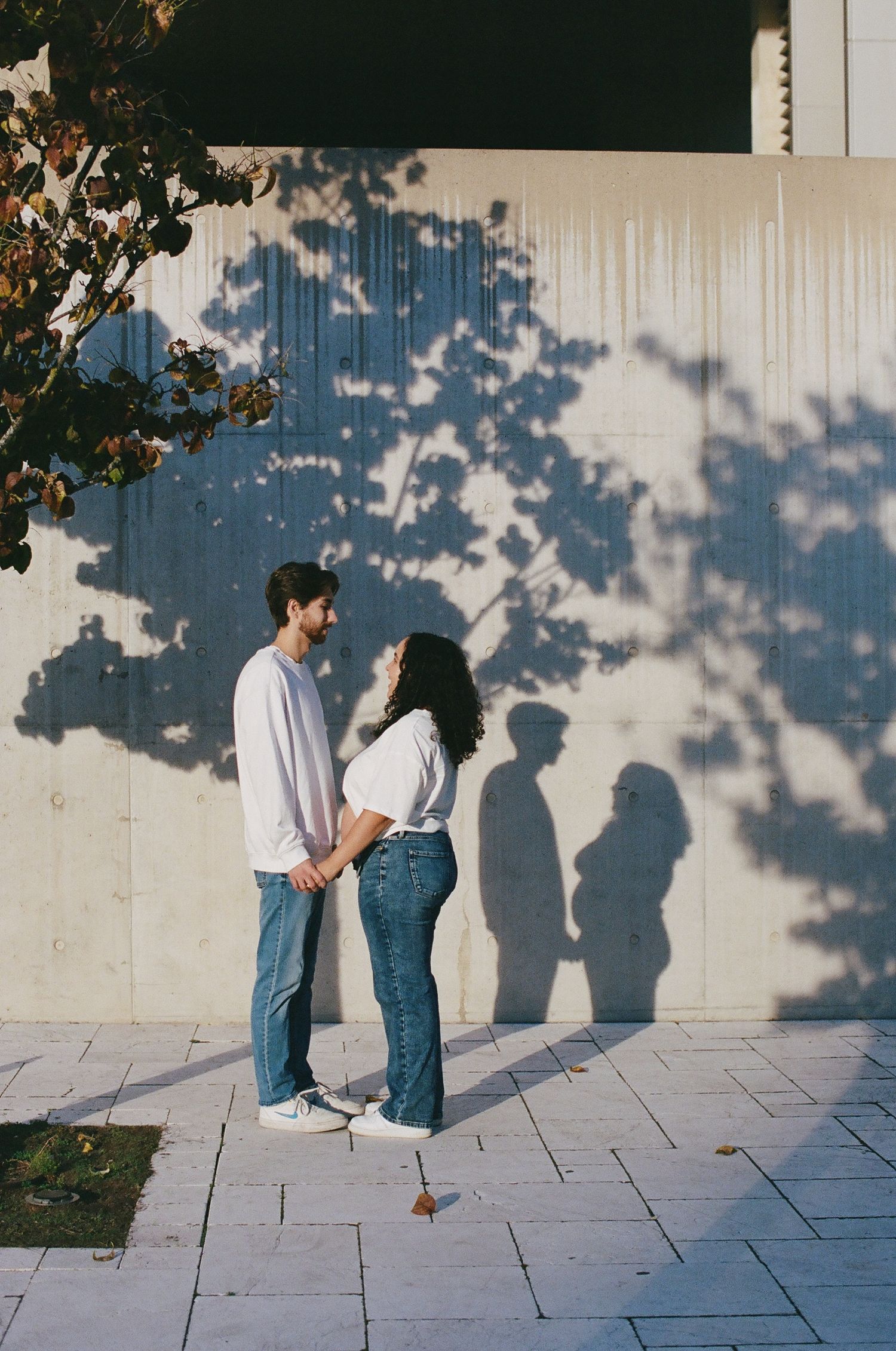 Tree shadows cast on a white wall as a couple poses during an outdoor maternity session.