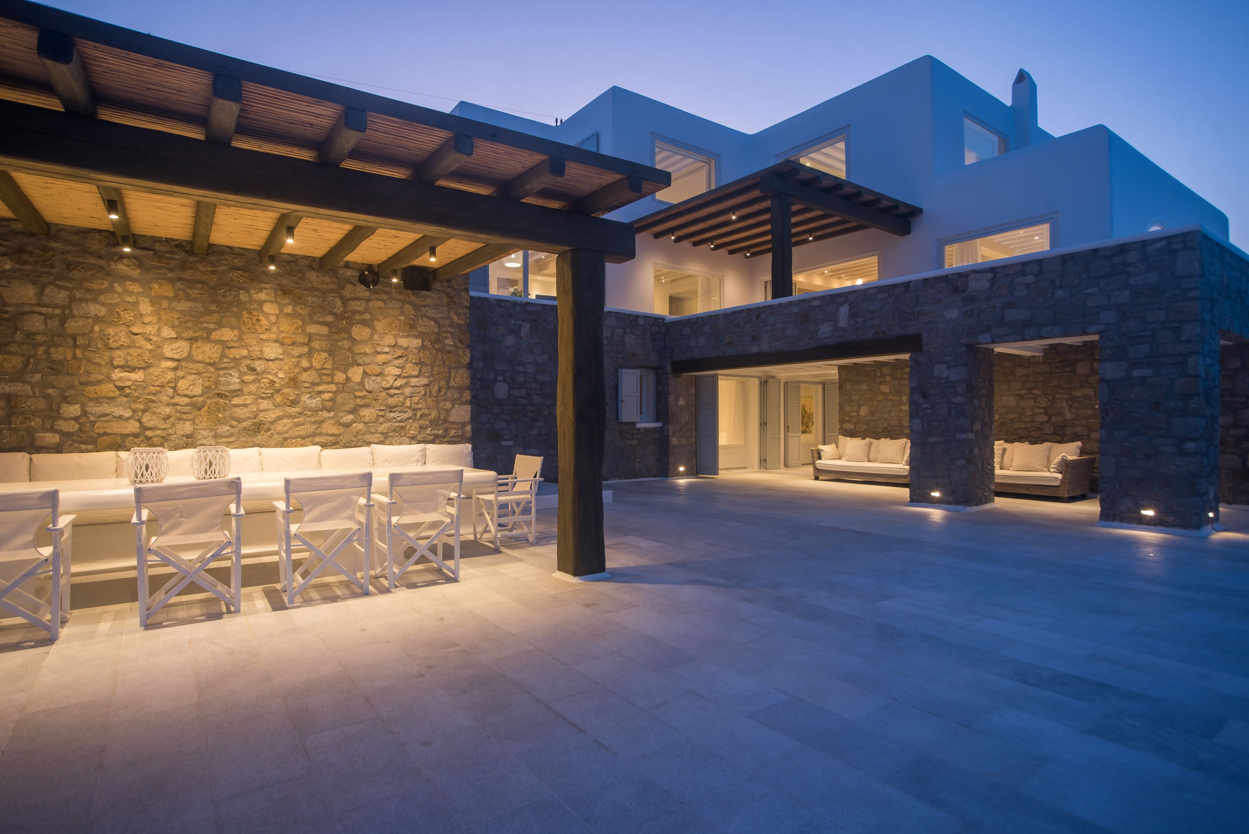 Modern luxury home exterior with stone walls and wooden pergola illuminated at dusk against blue sky.