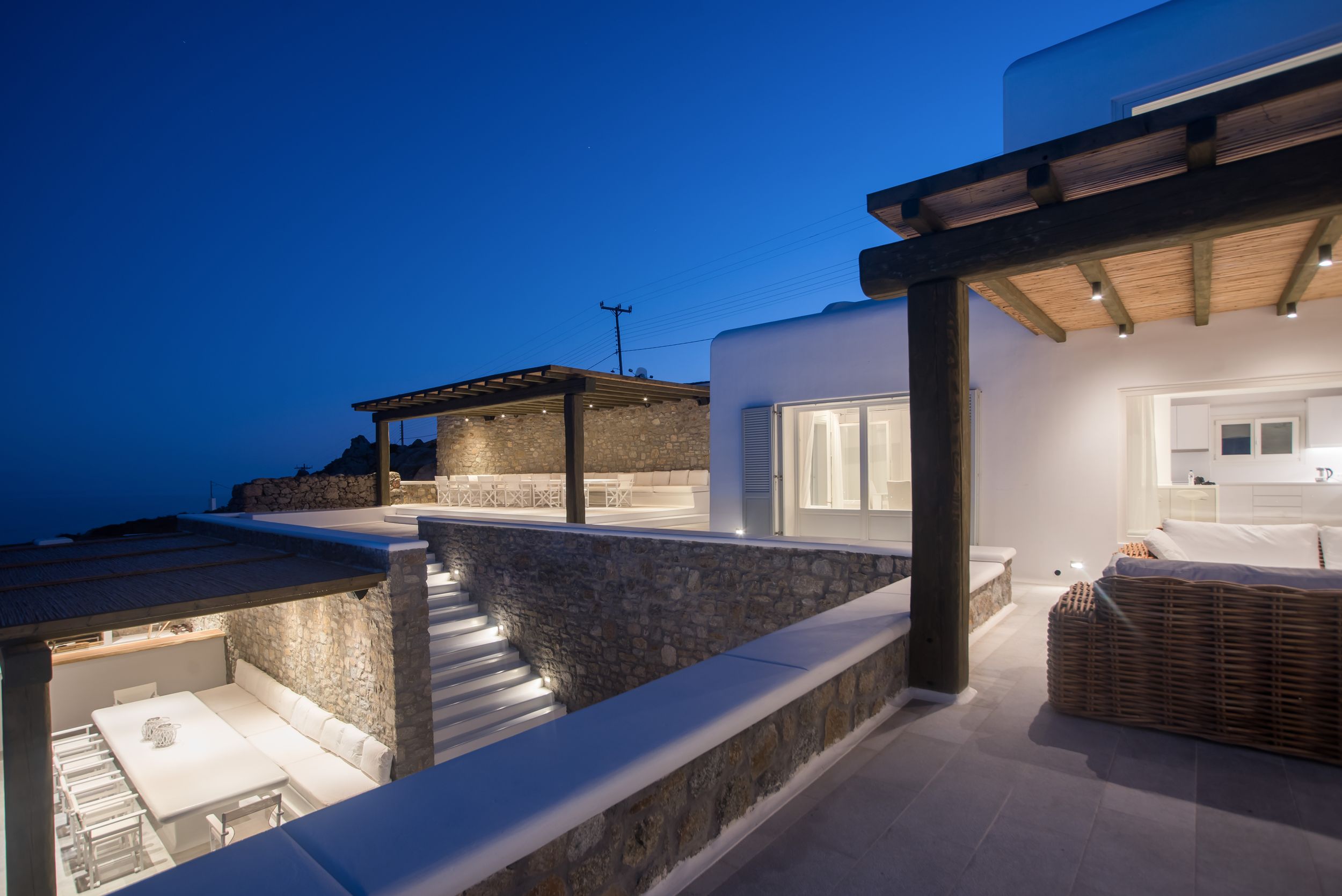Mediterranean villa terrace with white walls and stone steps illuminated at dusk against deep blue sky.Mykonos villa
