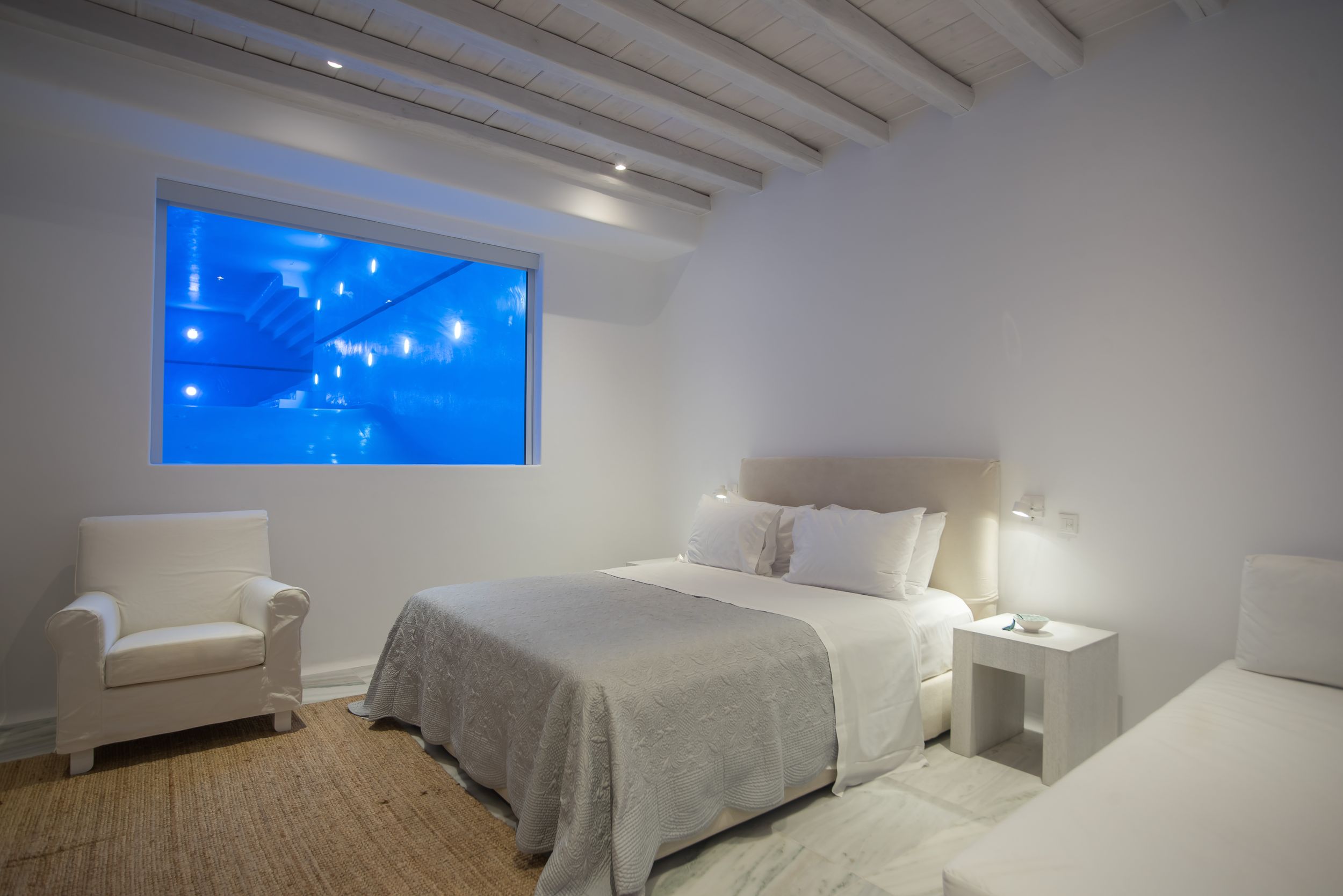 Modern white bedroom with glowing blue aquarium window display, white furniture, and exposed ceiling beams.
