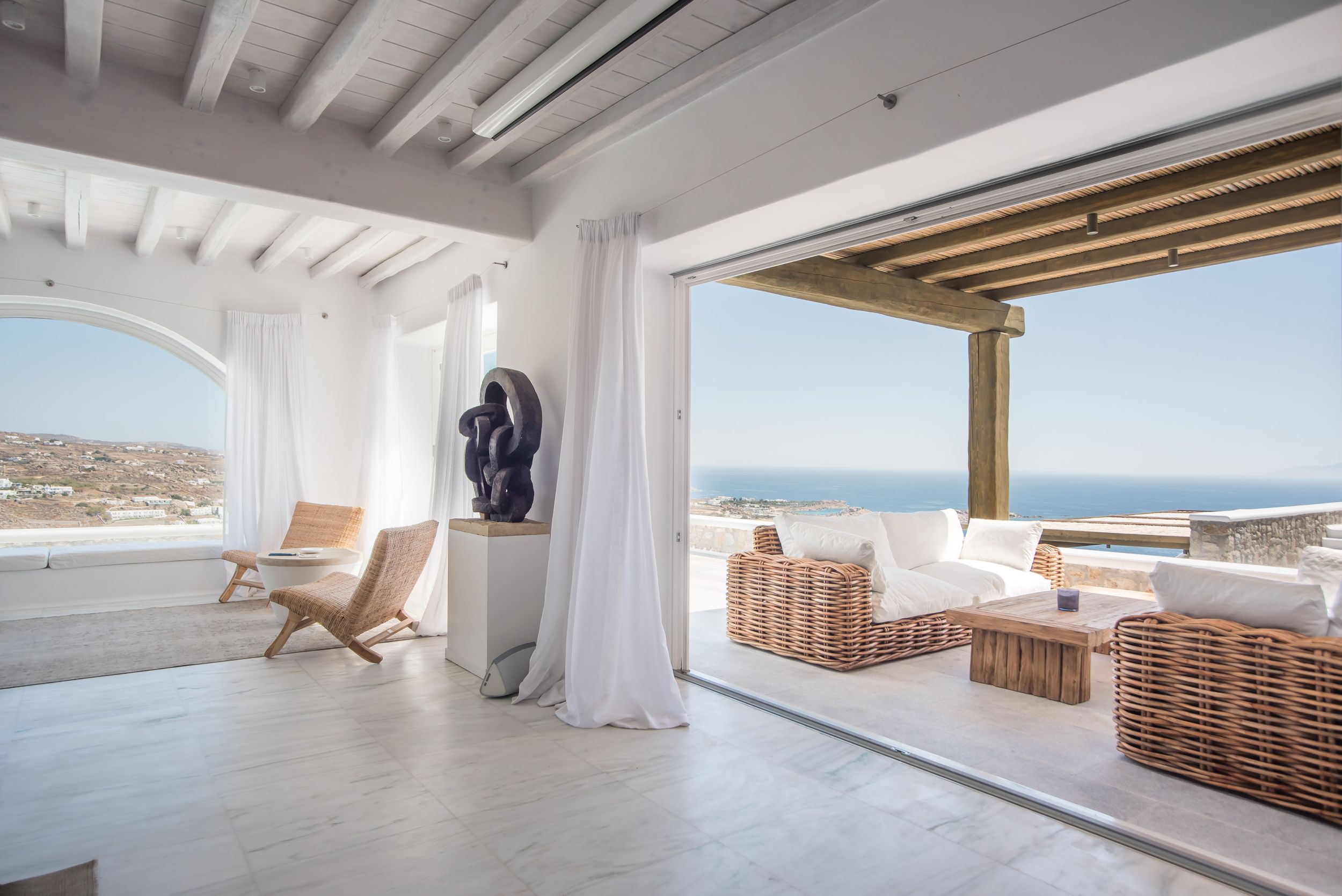 Luxurious Mediterranean villa interior with white walls and ocean view terrace featuring wicker furniture and natural decor.