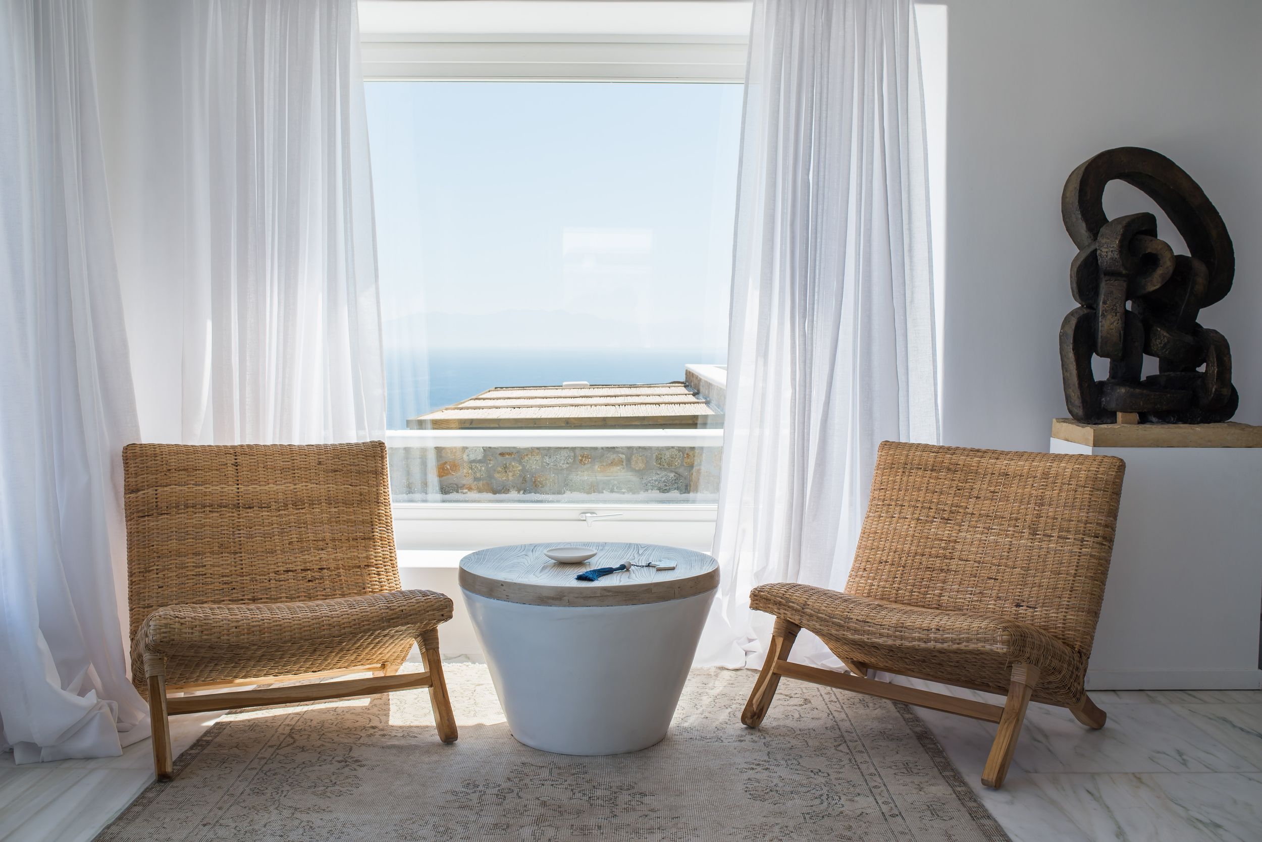 Modern coastal living room with wicker chairs and white drum table overlooking ocean view through sheer curtained window.