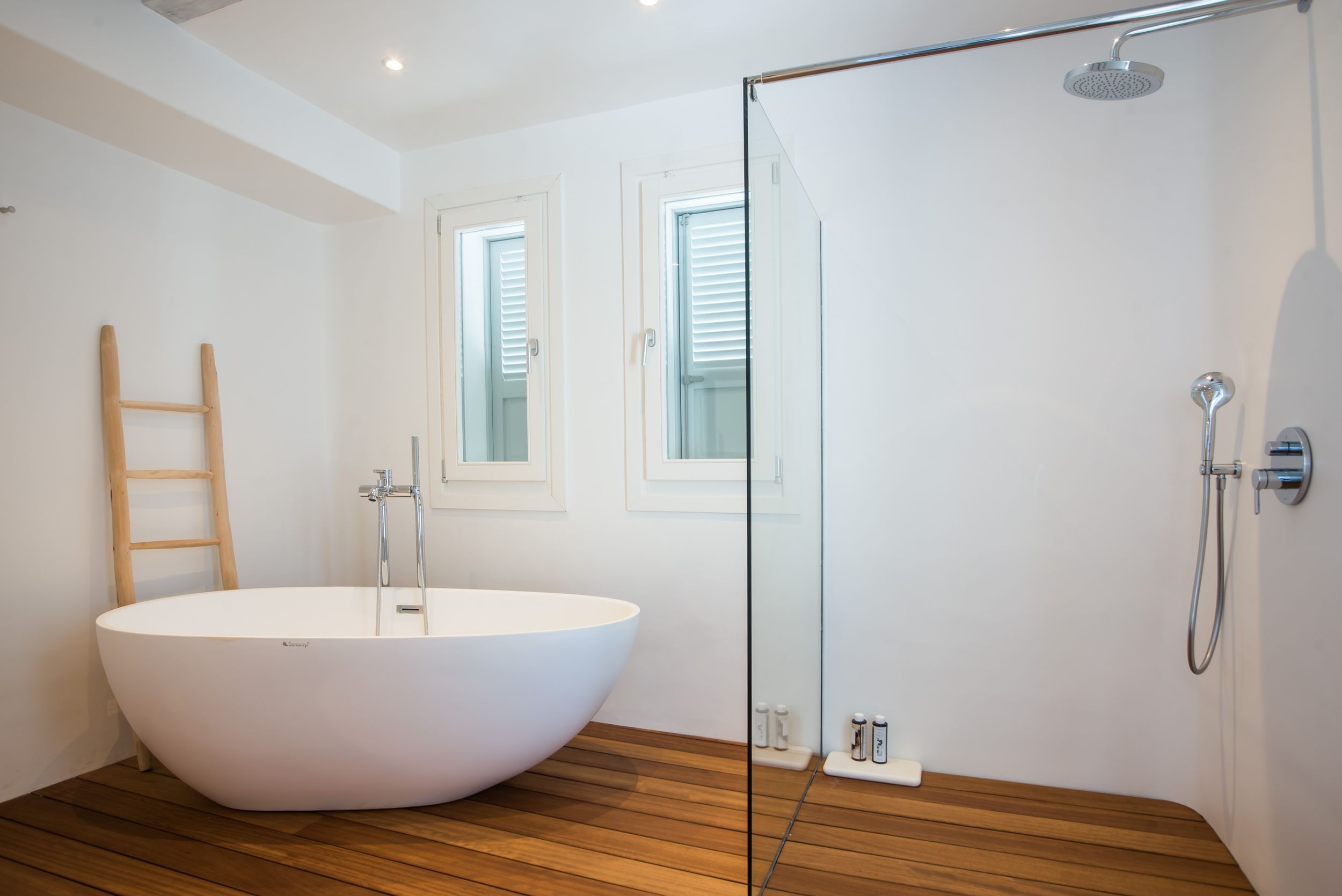 Modern white bathroom interior featuring a freestanding oval bathtub, wooden floor and glass shower enclosure.