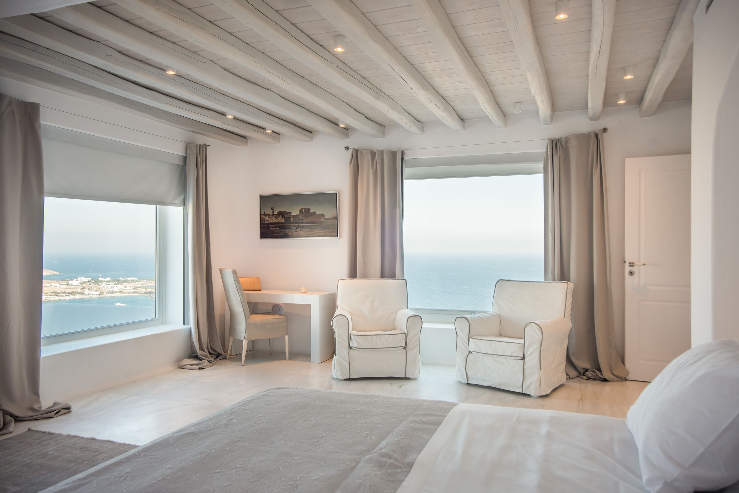 Luxurious coastal bedroom with white furniture and floor-to-ceiling windows offering stunning ocean views.Mykonos villa