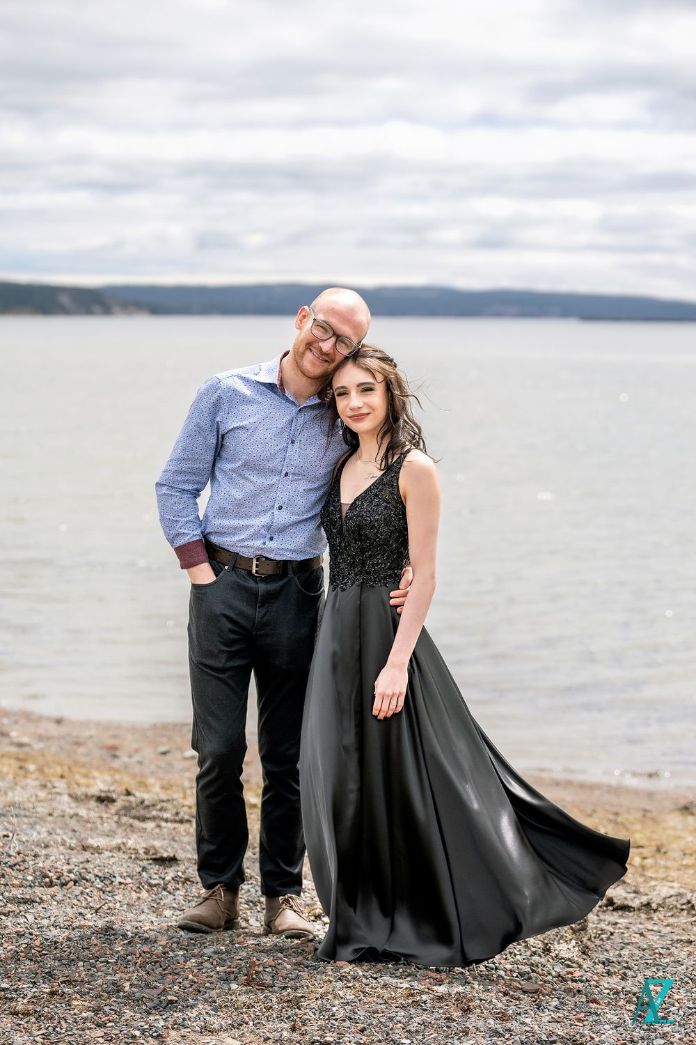 A couple poses together on a rocky shoreline in formal attire.