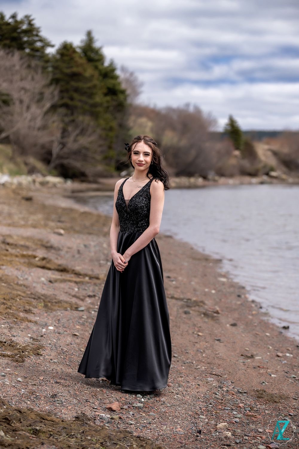 A woman in a flowing black gown poses on a rocky beach with trees in the background.