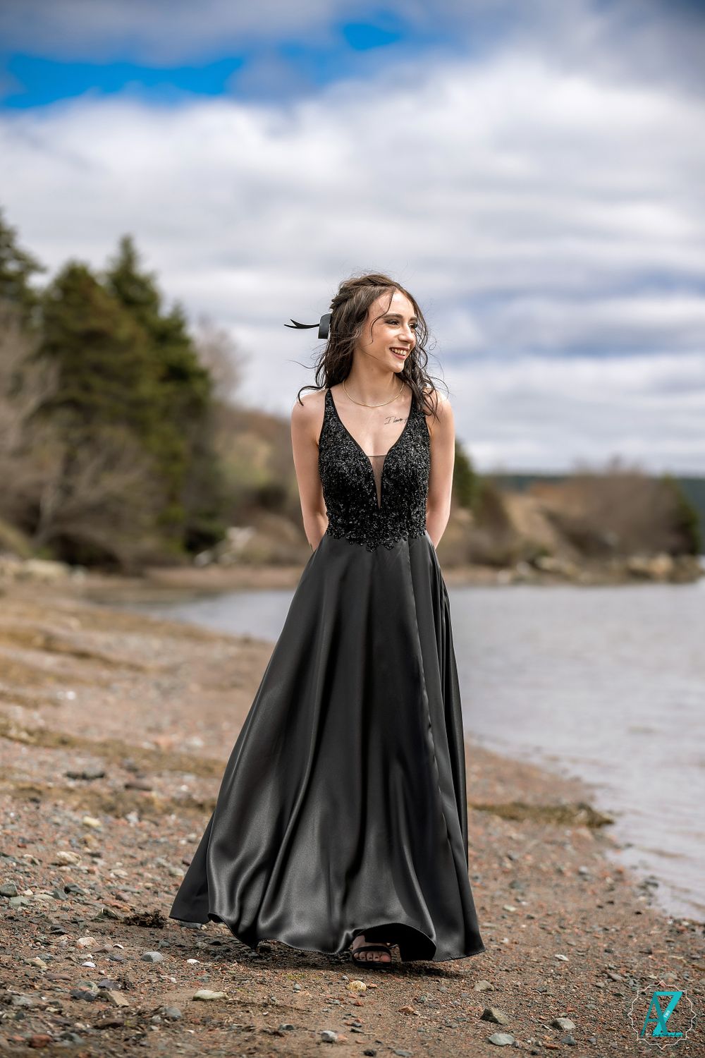 A woman in an elegant black formal gown poses on a rocky shoreline against a cloudy sky and water backdrop.