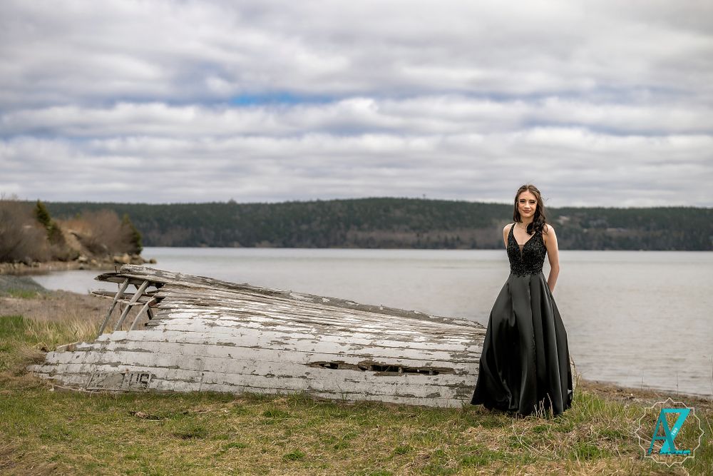 An elegant black gown contrasts beautifully against a natural waterfront landscape with driftwood and cloudy skies.