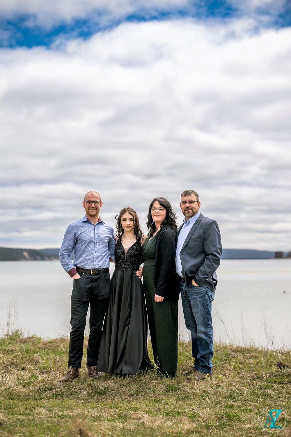 A group of four people pose together on a grassy cliff overlooking a scenic waterfront.