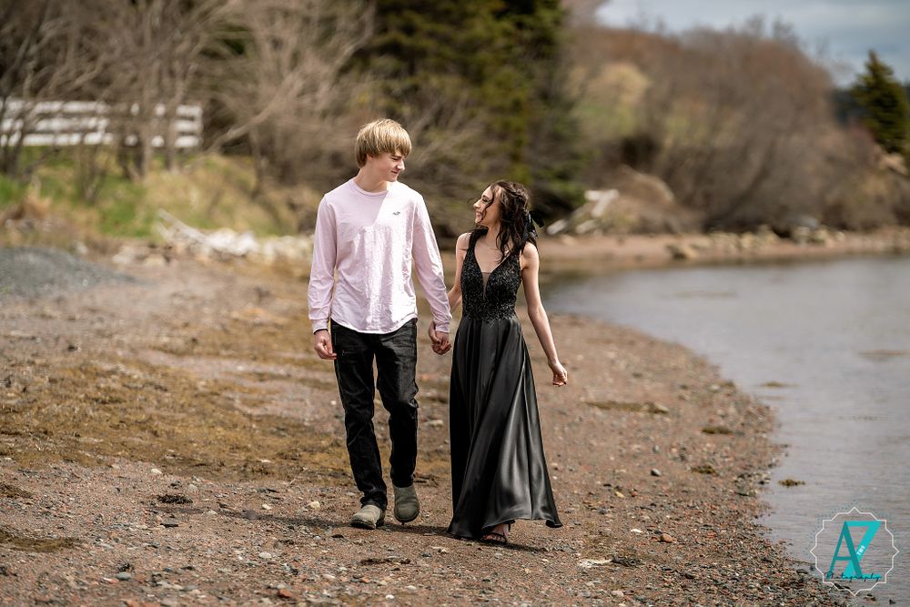 A couple walks hand in hand along a rocky beach shore wearing formal attire.