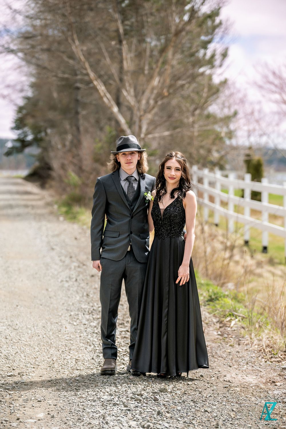 A formal couple poses on a gravel path lined with white fencing and trees.