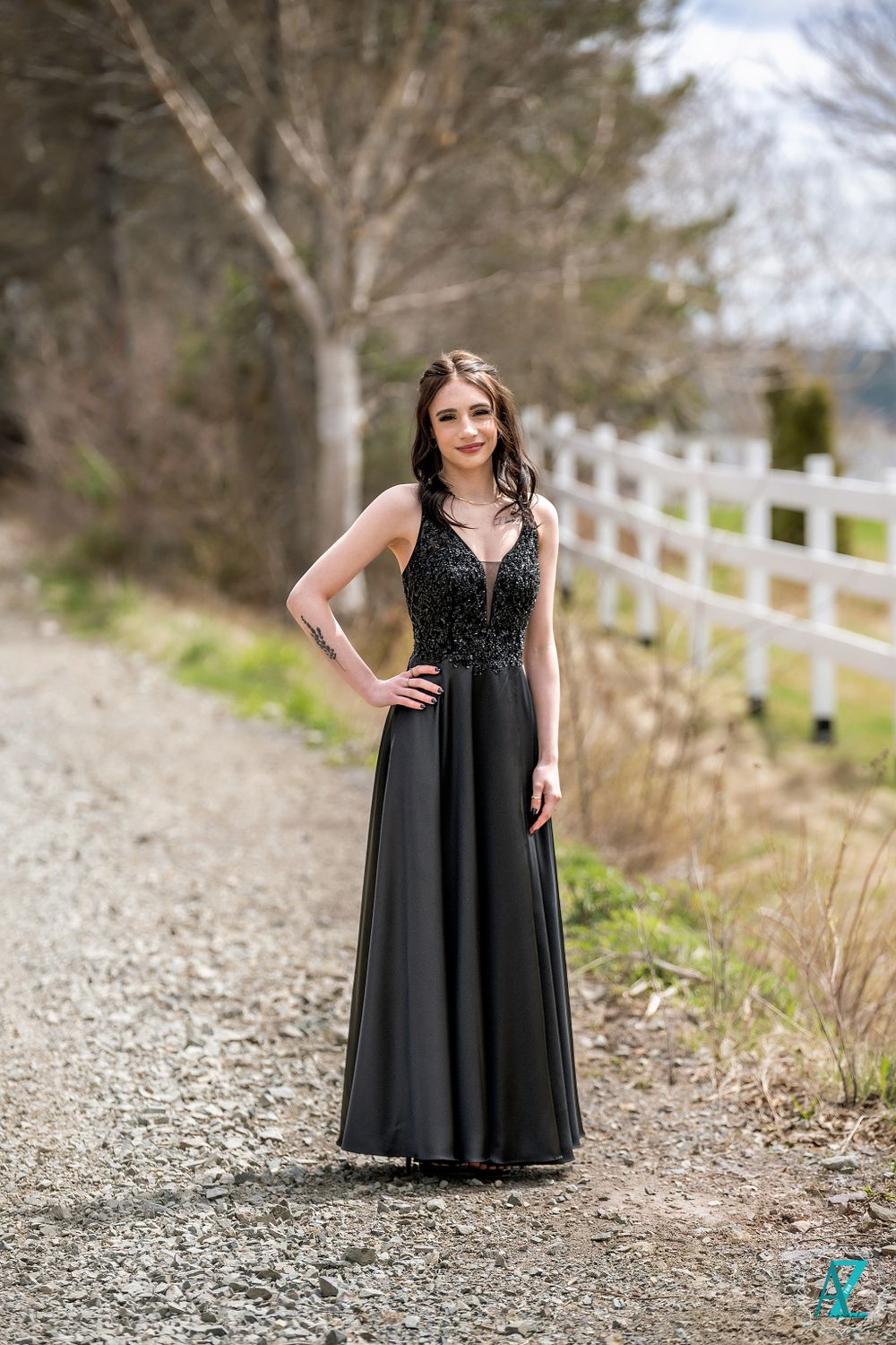 A formal black gown shimmers in natural light on a rustic gravel path with white fencing.
