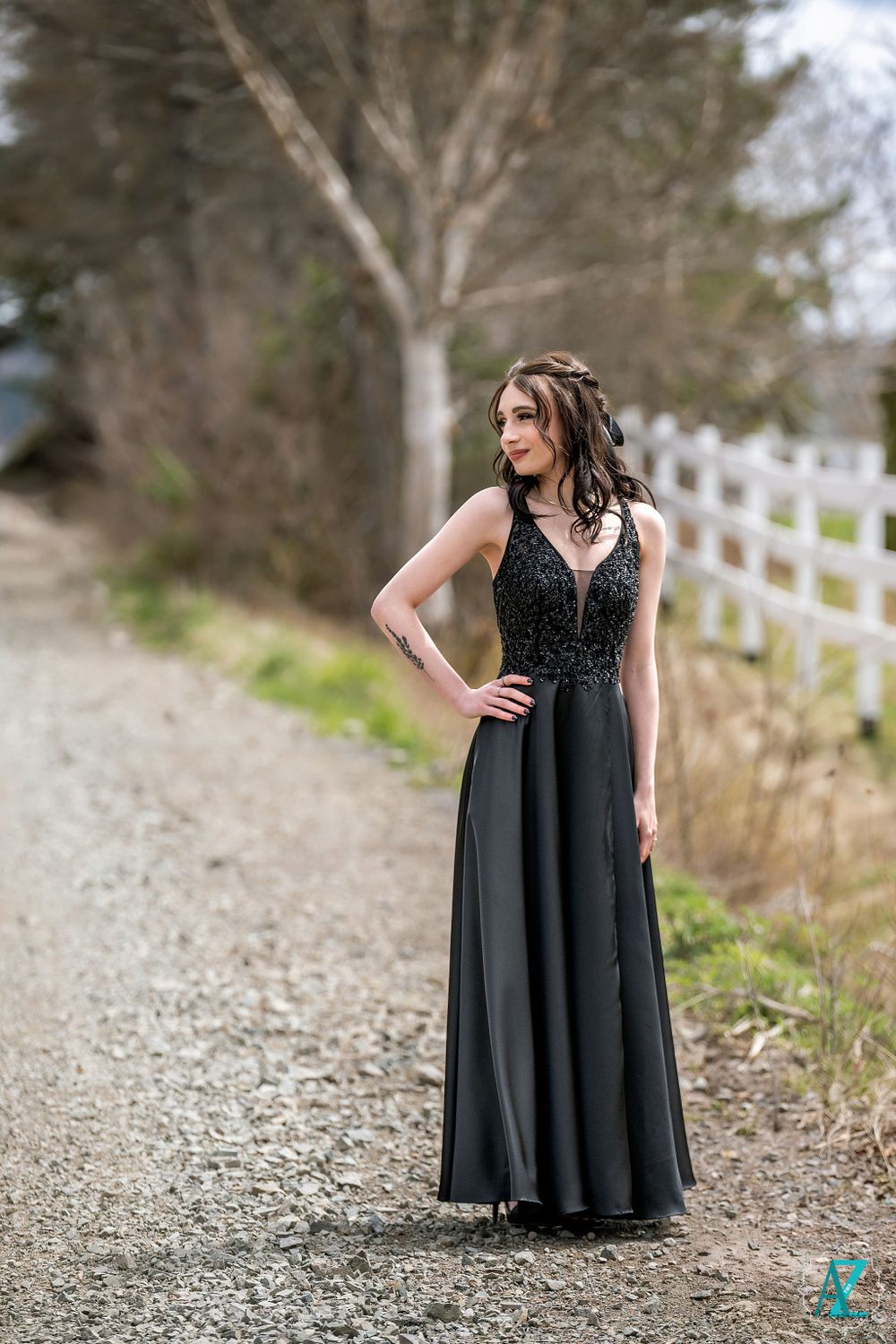 An elegant black evening gown catches light on a country path with white fence backdrop.