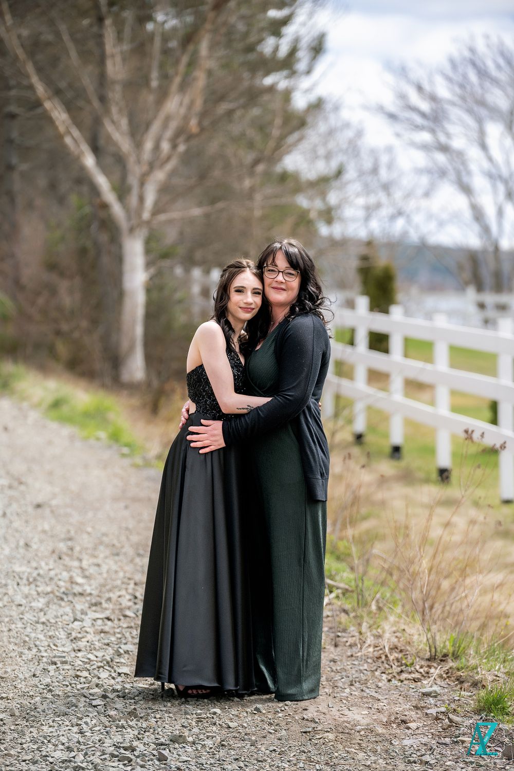 Two people embrace warmly on a gravel path with white fence and trees in background.