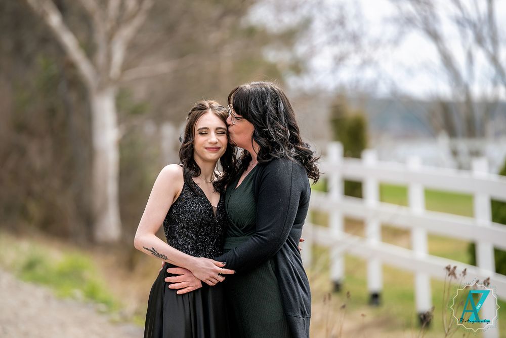 Two people in black dresses embrace on a scenic country path with a white fence in the background.