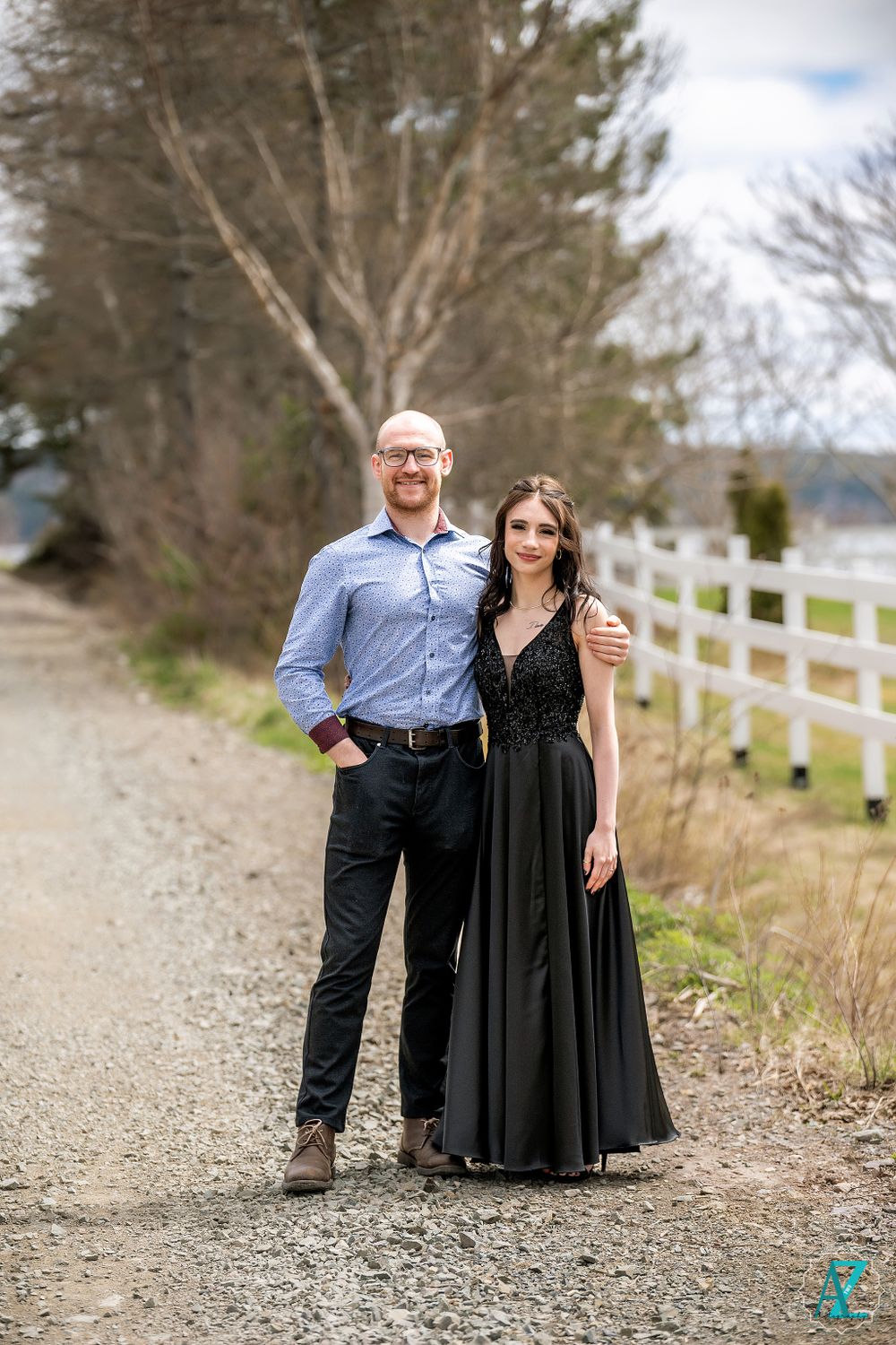 A couple poses on a gravel path, with the person wearing a formal black gown and the other in business attire.