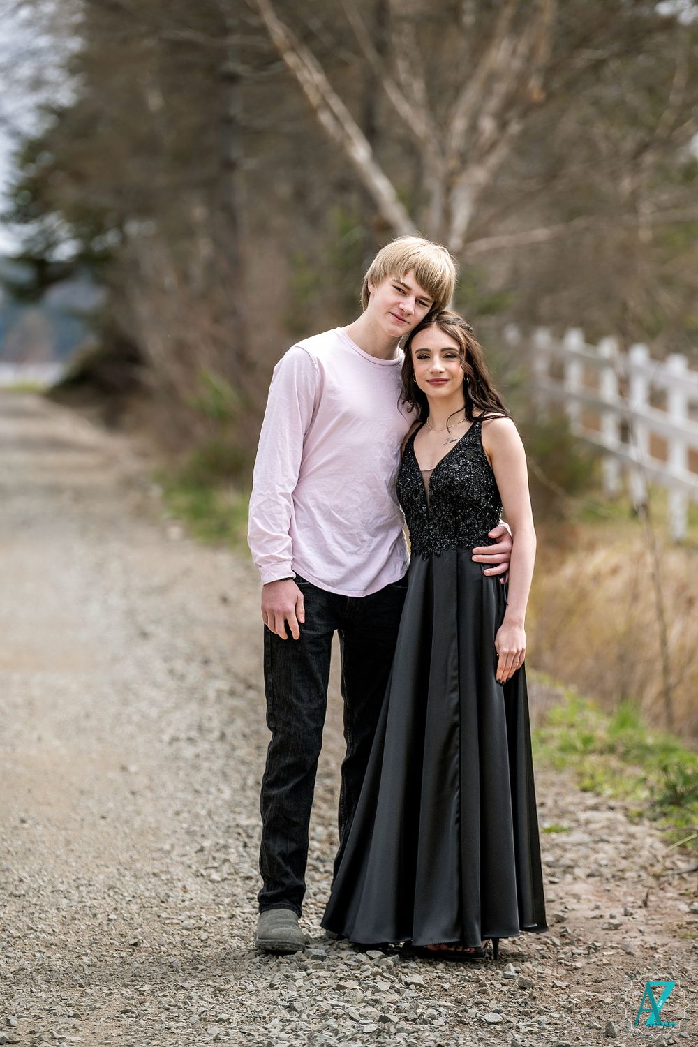 A couple poses together on a gravel road, one in a black gown and the other in a light pink shirt.