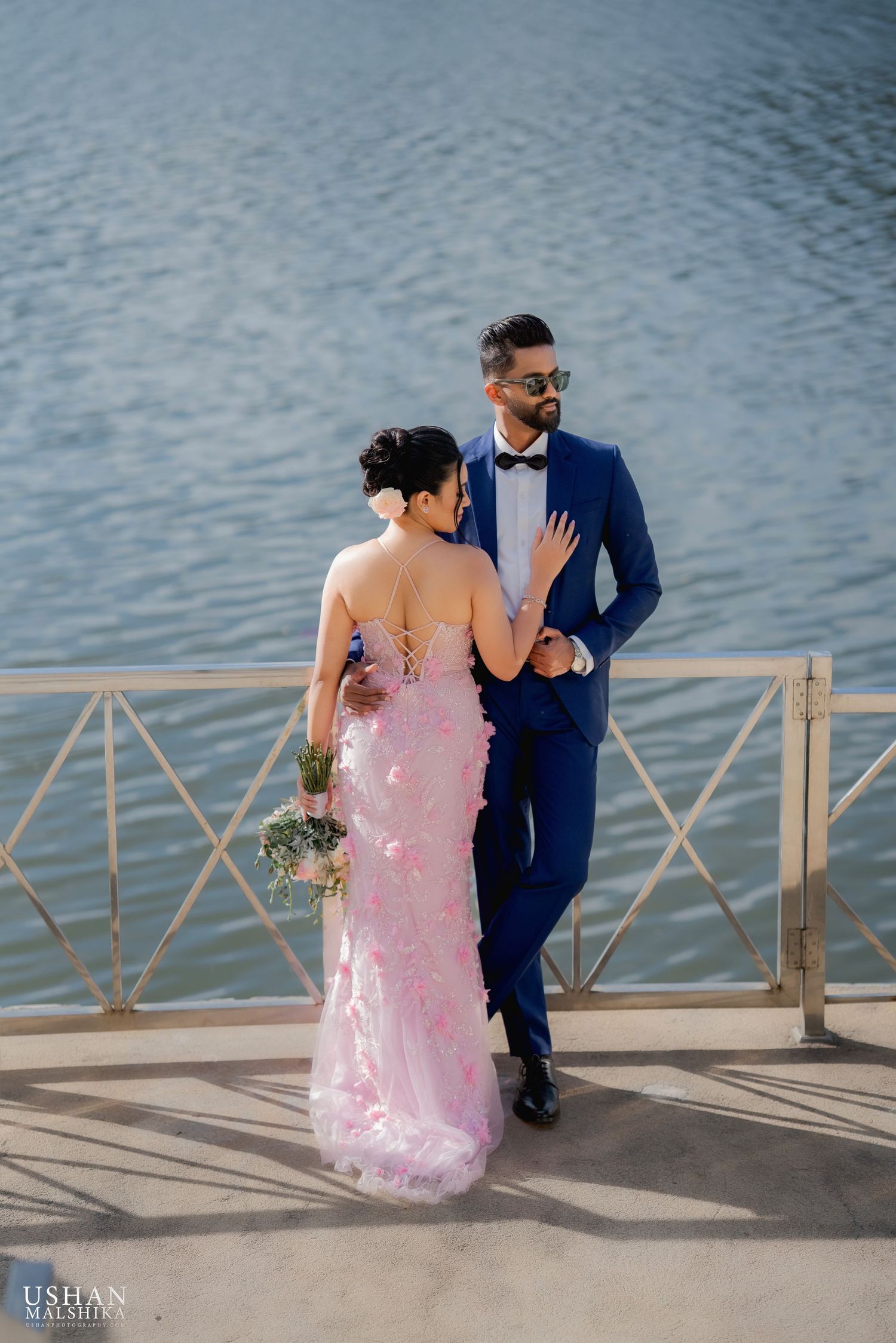Couple in formal attire posing on waterfront deck in romantic stance.