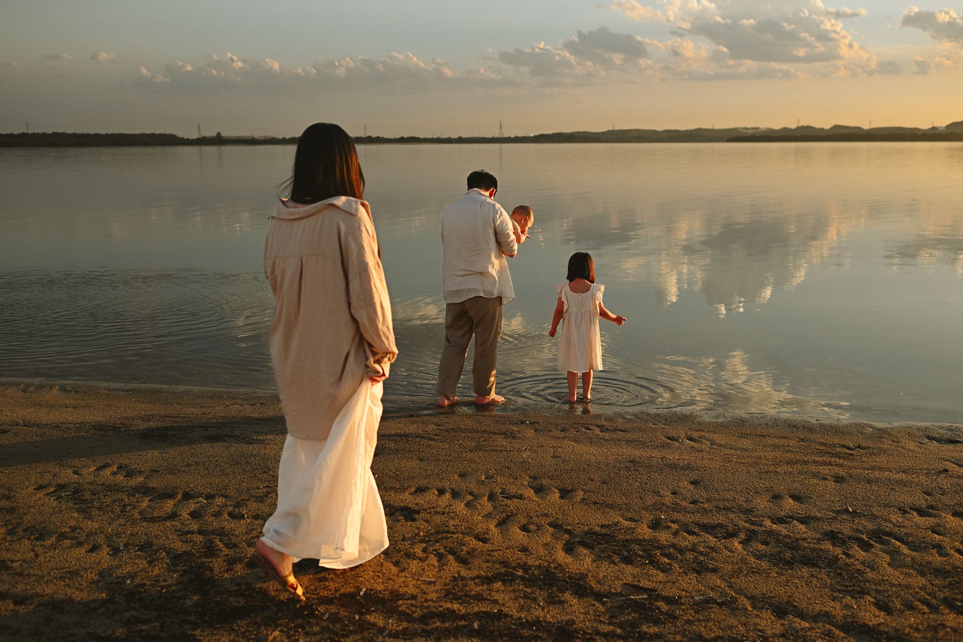A group in white clothing stands at the beach during a golden sunset, creating silhouettes against the peaceful water.
