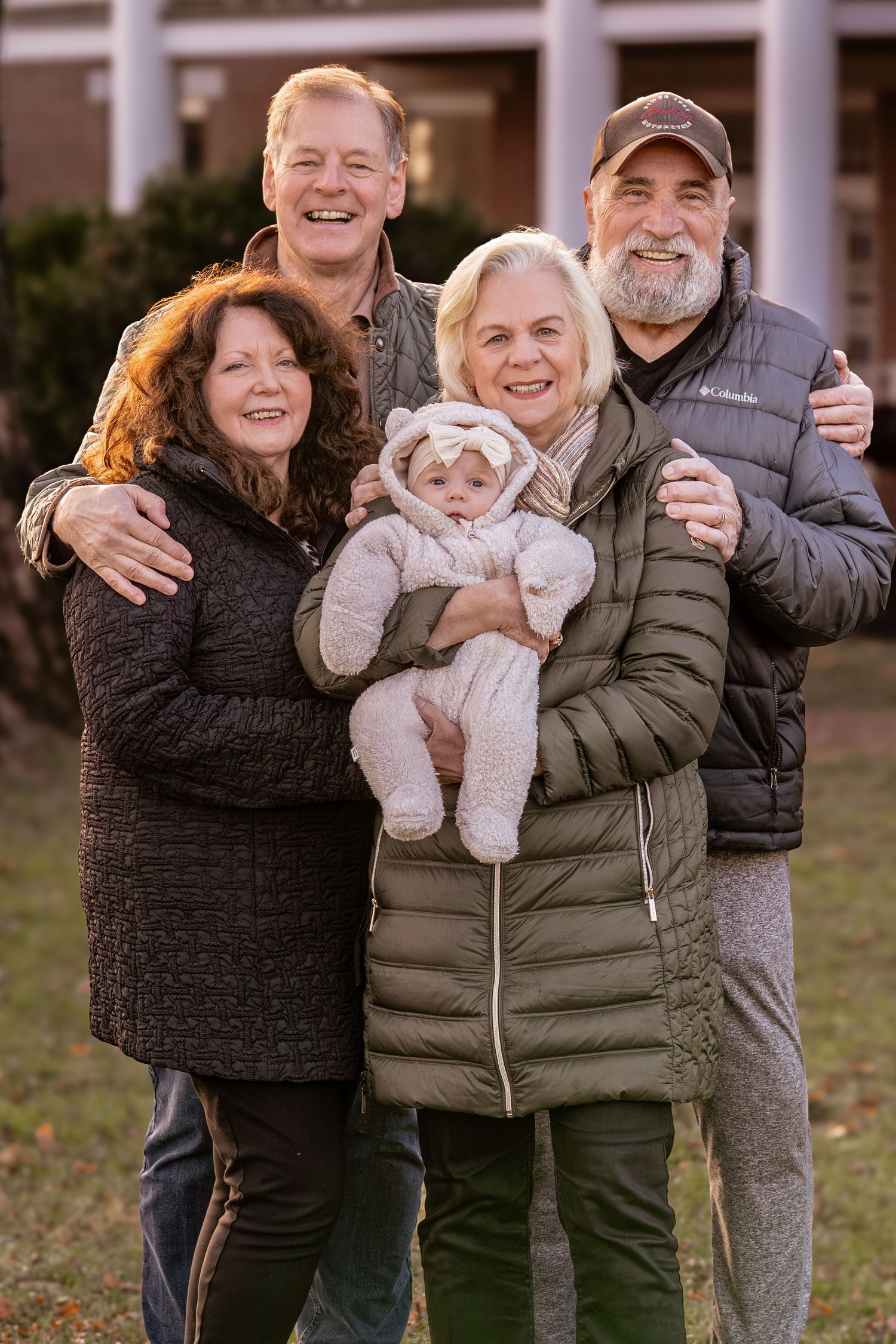 A group of people outdoors pose together in winter coats while holding a small baby and smiling at the camera.